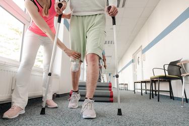 Unterkörper von männlichem Patient in der Reha an Krücken und Physiotherapeutin beim Ausführen von Gehübungen 